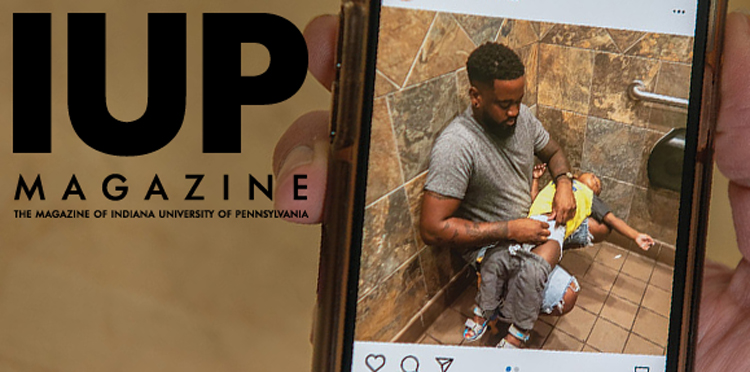 Closeup of a hand holding a mobile phone that displays an Instagram post with a photo of Donte Palmer squatting on the floor of a public restroom with his back against a wall as he changes a toddler’s diaper on his lap