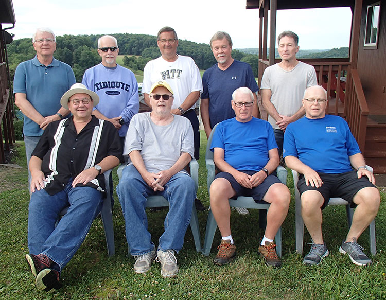 Nine men around 70 years old pose for a photo outdoors between two cabin-type structures. Four of them sit in plastic lawn chairs in front, and five stand behind them.