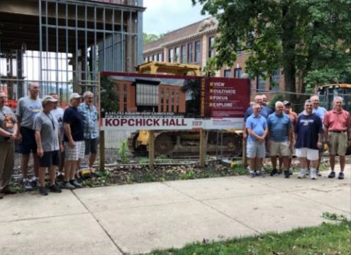 At least 14 men pose on either side of a sign about Kopchick Hall while the building, in an unfinished state, is visible behind them. McElhaney Hall is behind them on the right.