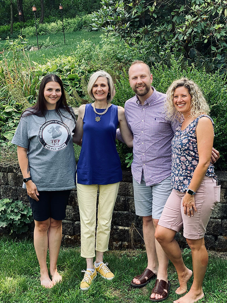 Three women and a man in casual clothing pose in a backyard-type setting with plants and bushes and a block wall behind them.