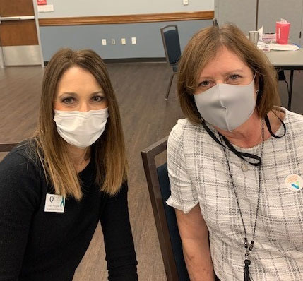 Two women wearing face coverings sit in a large, open room and face the camera