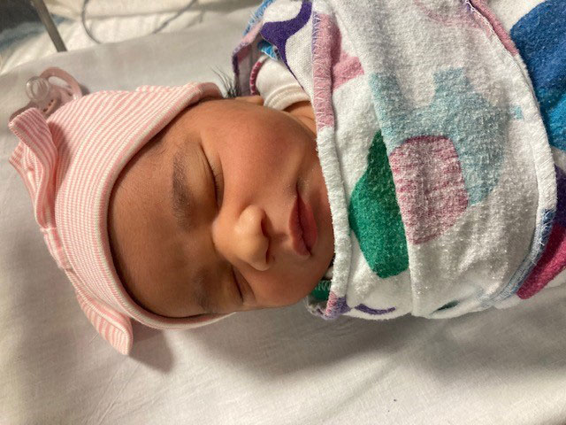 A baby wrapped in blankets and with a cap on her head has her eyes closed as she lies in a hospital bassinet.