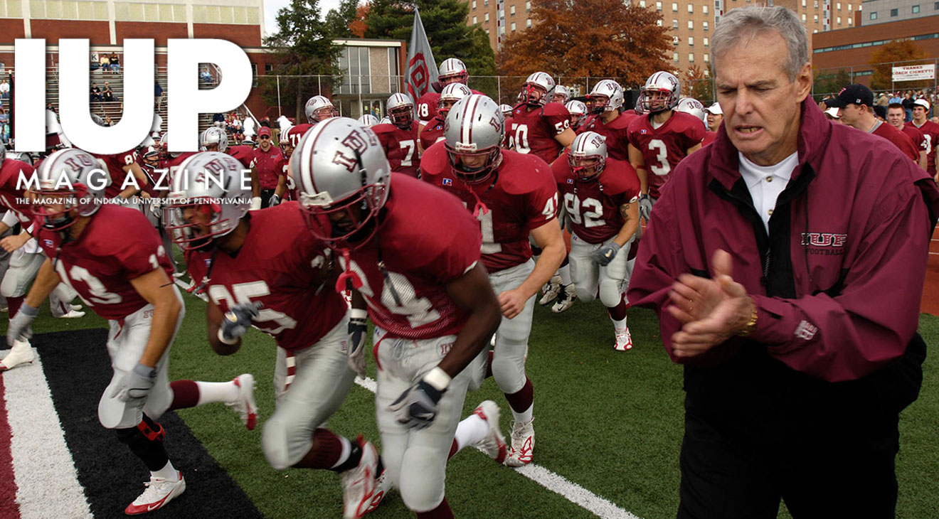 The IUP football team, in silver helmets, red jerseys, and white pants, runs onto a football field next to coach Frank Cignetti, who is clapping in a dark red IUP sweat suit.