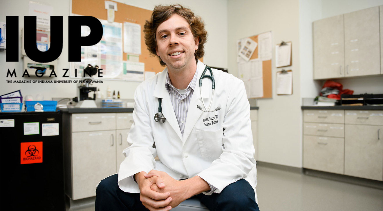 Joe Rocco wearing a buttoned, white lab coat over business casual clothing and a stethoscope around his neck sits on a chair in the middle of a medical exam room or lab with a microscope in the background. He smiles for the camera as he leans slightly forward with his forearms resting on his legs and his hands clasped in front of him.