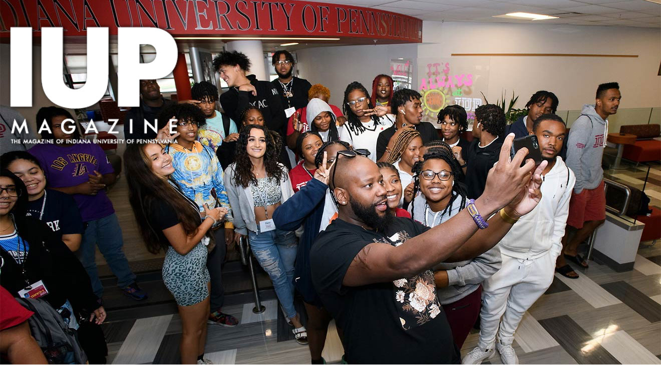 A young man with more than 20 younger people standing behind him holds up his phone facing himself and the crowd to take a selfie. They are in a large, lobby-type space with an “Indiana University of Pennsylvania” sign in red near the ceiling and a carpet with long rectangles in shades of gray.