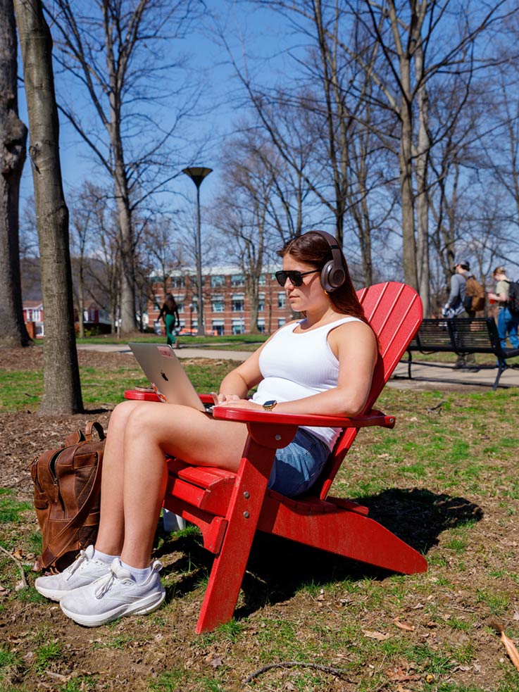 A student wearing sunglasses, blue shorts, and a white top wearing headphones sitting in a red Adirondack chair in the Oak Grove on a sunny day, with a laptop computer open in her lap