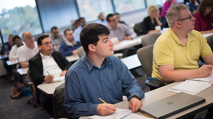 A male student is seated at a table in an MBA classroom with other students