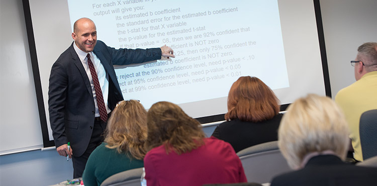 A professor stands in front of a projector screen, pointing something out to the MBA class A professor stands in front of a projector screen, pointing something out to the MBA class