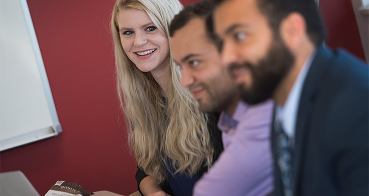 Three students in an MBA classroom, two male that are out of focus and a female student that is looking towards the camera