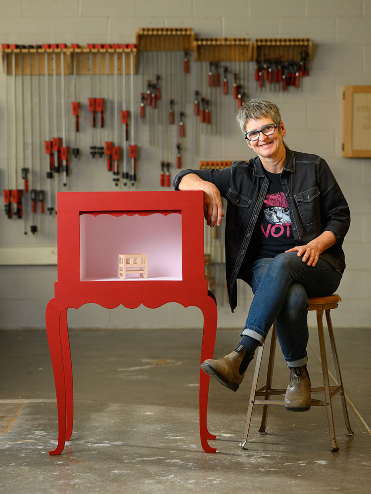 BA Harrington poses in the studio next to a case holding a small artwork