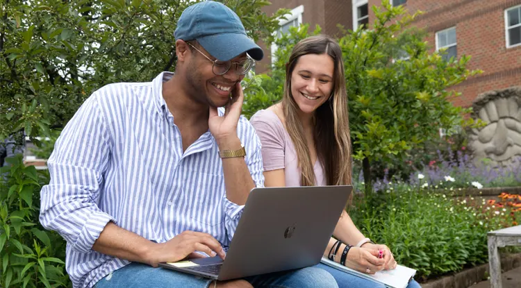 Two students smiling and looking at a laptop screen while sitting outside