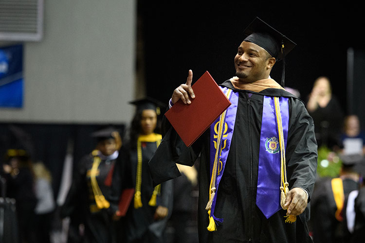 A young man in a graduation cap and gown with a purple sash with fraternity letters and at least three yellow or gold cords around his neck walks in an arena setting with two others in graduation attire behind him. The man is smiling and holding up a red diploma cover in his right hand and pointing with the same hand.