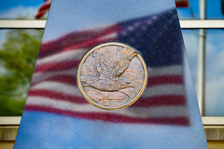 Detail of the flat face of a bluish, triangular-shaped monument that has at its center the sculpted image of a flying eagle in a bronze circle, surrounded by the reflection of a flying American flag on the monument’s surface. Behind the monument are highly reflective windows of a building showing a tree and a flagpole.