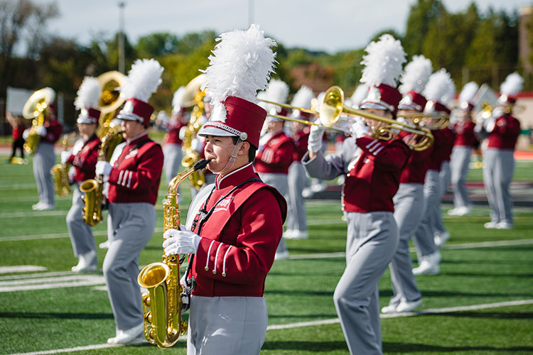 Members of the marching band in red jackets with the IUP logo, gray pants, white gloves, and red hats with white plumes perform on the football field on a sunny day. Saxophone players are in the foreground, with trombonists behind them and tubas in the background.