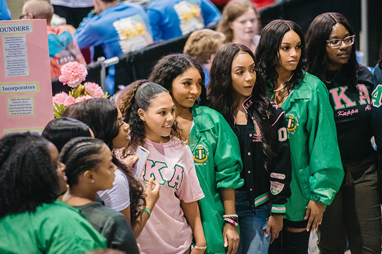 Nine young women wearing green, pink, or black clothing with sorority letters stand in a row and look to their left as if posing for a photograph in a crowded, arena-type setting.