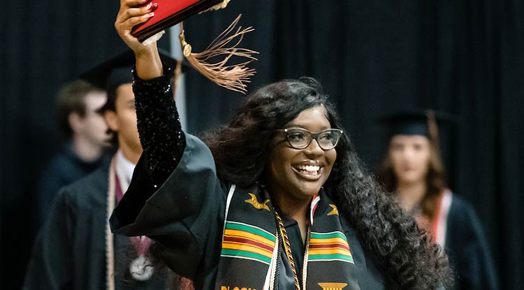 Graduate smiling on stage and holding aloft her diploma case 