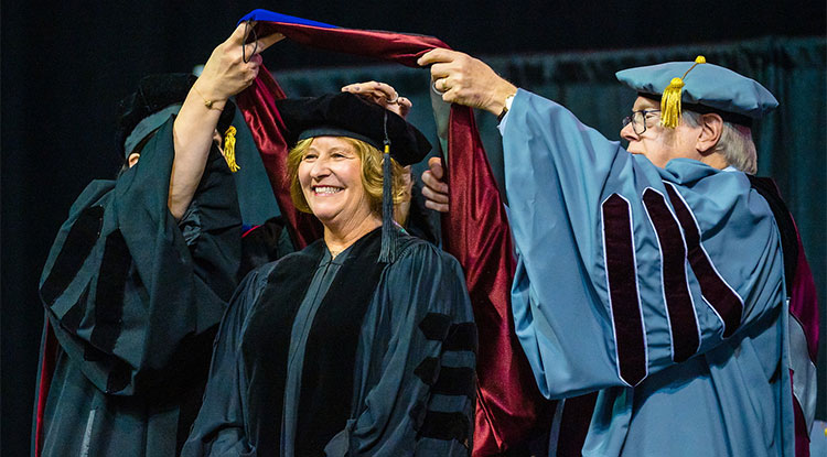 A woman in a black cap and graduation gown with black velvet down the front smiles as two people hold a burgundy sash over her head.