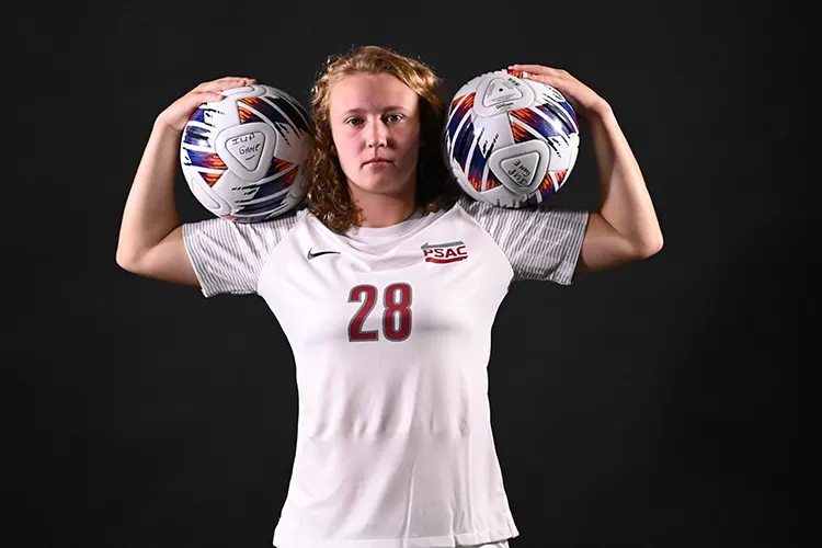 Hannah Scardina facing the camera while holding to soccer balls on her shoulders