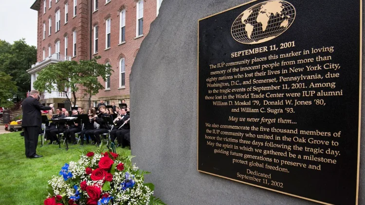A closeup of the IUP 9/11 memorial stone in the Oak Grove.  A bouquet of flowers sit at the base while an orchestra performs in the background in front of Sutton Hall