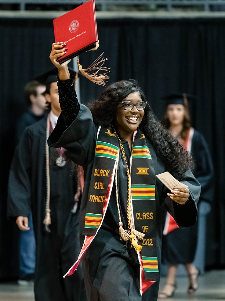 Graduate smiling on stage and holding aloft her diploma case 