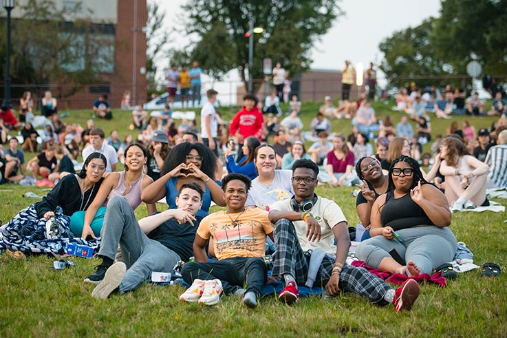 A group of IUP students sitting on the lawn and smiling and posing for the camera, with many other students behind them. 