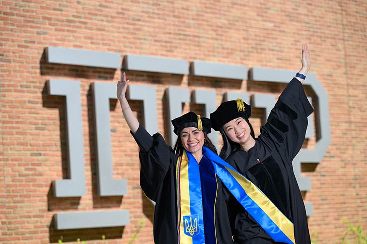 Two graduates pose for a photo, dressed in their caps and gowns, in front of the IUP logo