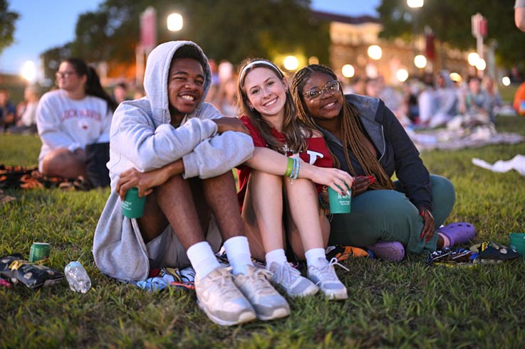 Three students sitting on the grass of Foster Lawn in the evening, waiting for the outdoor movie to start