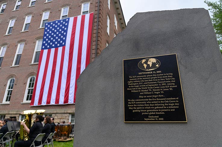 A plaque honoring the victims of September 11, 2001, mounted on a block of granite in the IUP Oak Grove, behind which are seated musicians and an American flag hanging vertically from Sutton Hall.