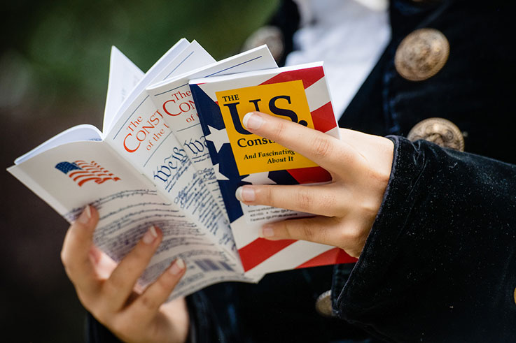 Hands of a person in 18th-century clothing holding pamphlets of the US Constitution