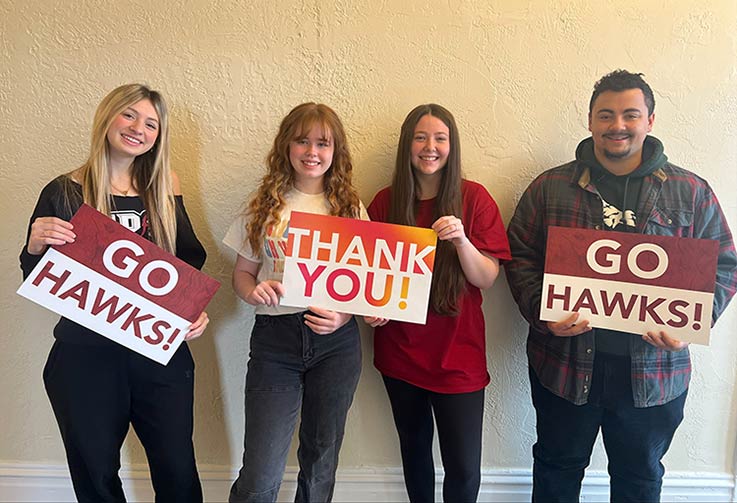 Four smiling students holding signs of saying Thank You and Go Hawks.