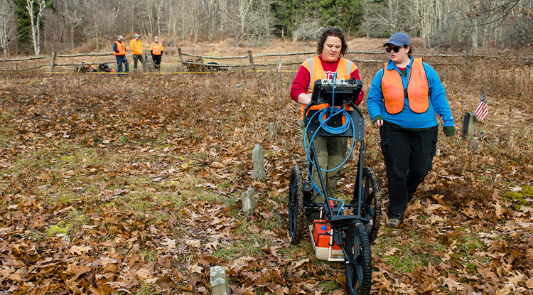 One woman pushes a ground penetrating radar device while another walks alongside her through an old cemetery with sparse, rudimentary gravestones in the fall.