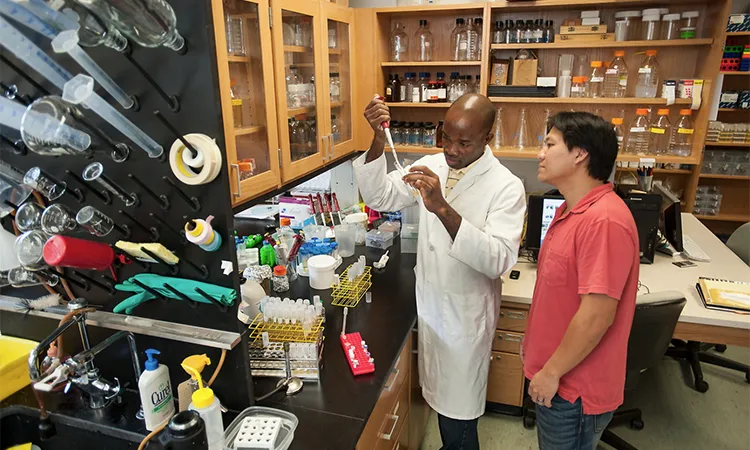 a man in a white lab coat adds a chemical to a vial while a man in a salmon colored shirt looks over his shoulder