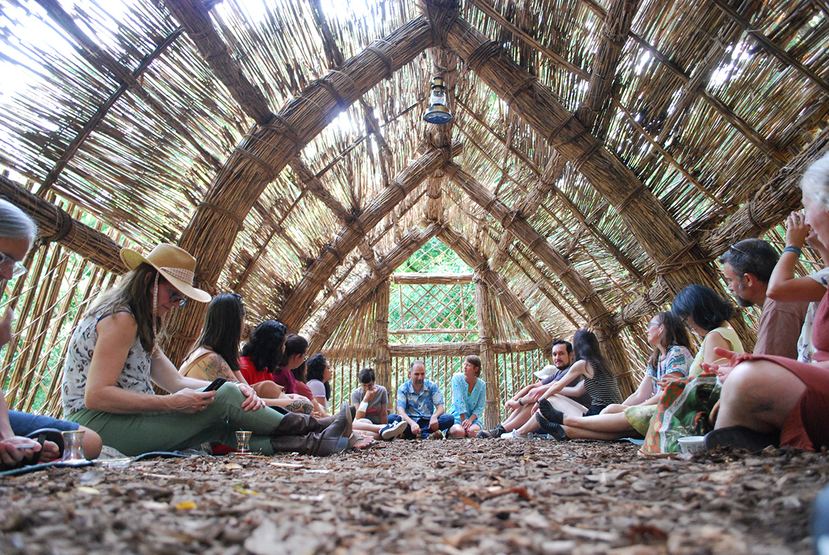 Several people sit on the ground inside a structure made of reed grasses as light streams through the ceiling