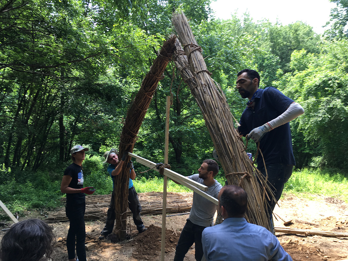 Five people work on the start of a structure, tying together two columns made of reed grasses to meet at the top and form an arch, while using wooden planks to measure the space in between