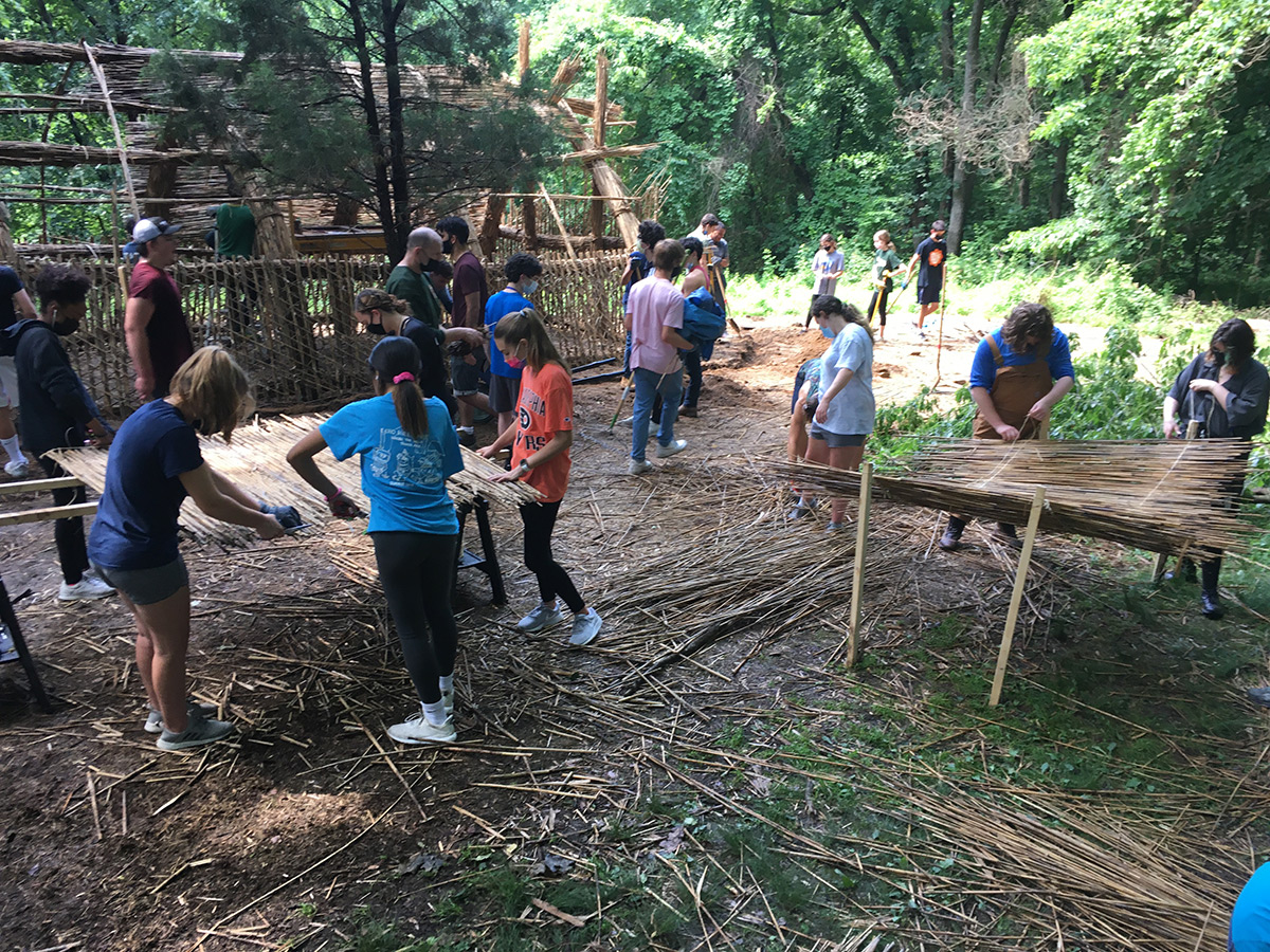 Several volunteers move in the wooded area around where the guesthouse is being built, while two small groups assemble sheets of tied-together reed grasses for thatching the structure’s exterior