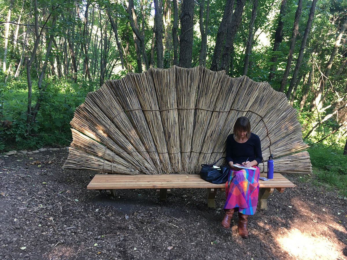 Artist Sarah Kavage writes in a notebook while sitting in the woods on a bench with a wooden seat and a back made of columns of reed grasses in a fan-like formation