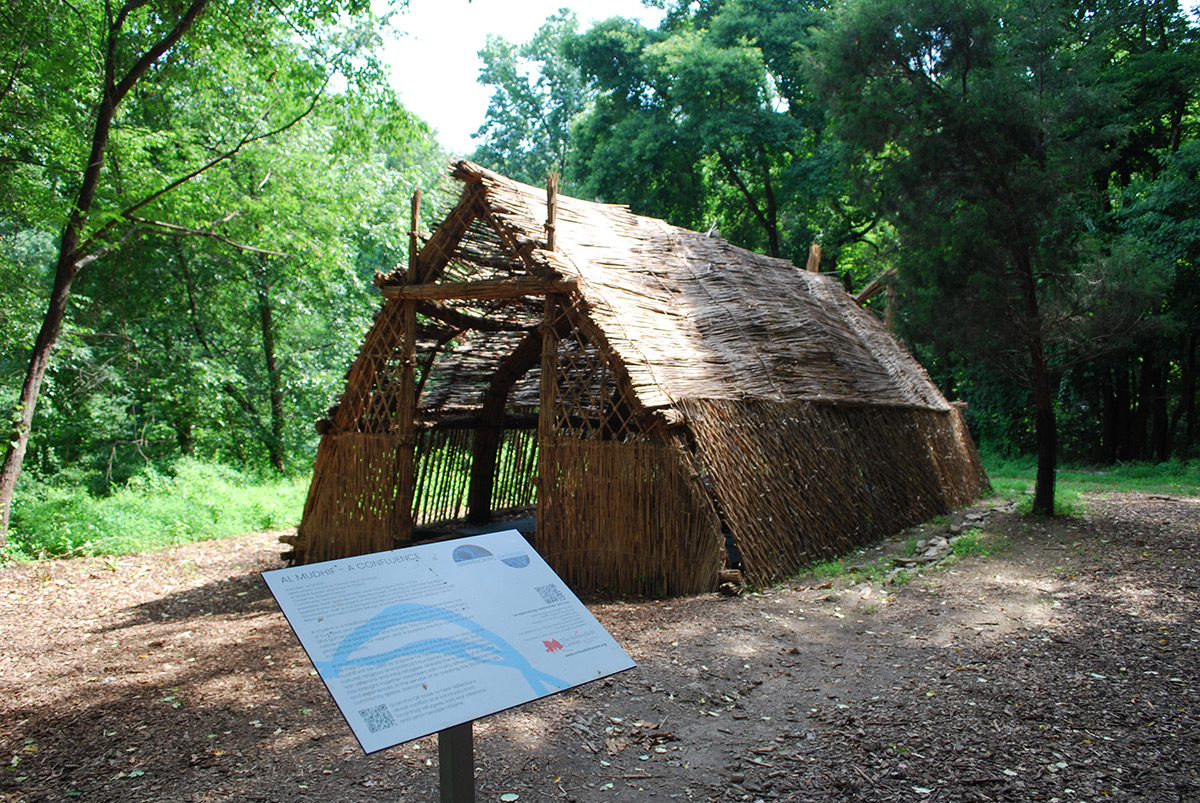 A view of the exterior of a completed structure made of reed grasses, with an opening for the doorway and a sign on a stand in front of the structure with a long description of what it is