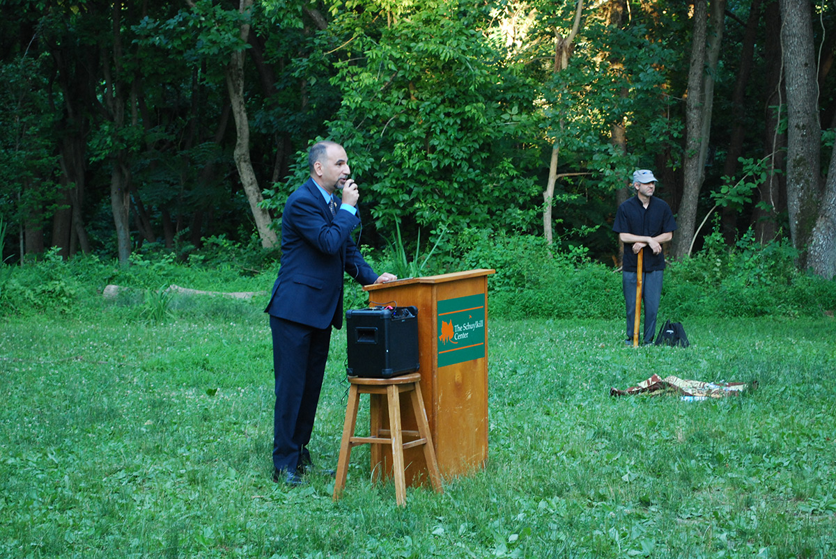 Yaroub Al-Obaidi speaks at a podium, with another man in the background, in a grassy area with woods in the background