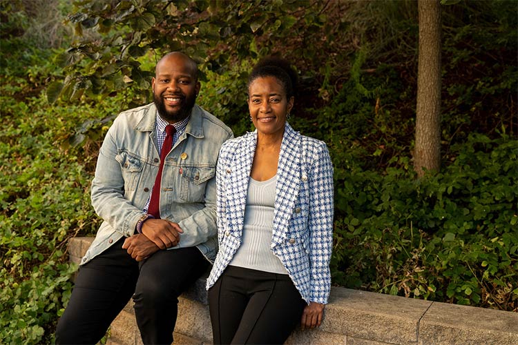 A man sits and a woman leans on a short gray-stone wall with lots of shrubbery around it as they look at the camera and smile. The man is wearing a jean jacket with a shirt and tie under it. The woman is wearing a jacket with a check pattern over a light sweater.