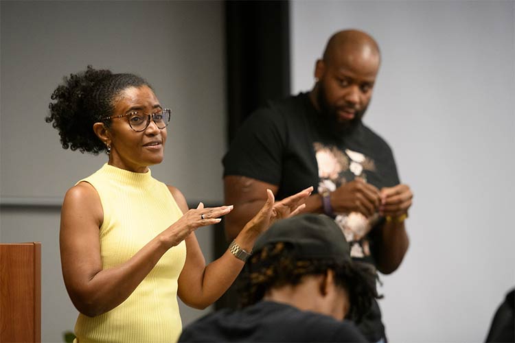 A woman wearing a sleeveless yellow mock turtleneck and her hair in a ponytail motions with her hands as she talks to people who are seated in a room, and a man who is also standing at the front of the room, in the background, watches her talk.