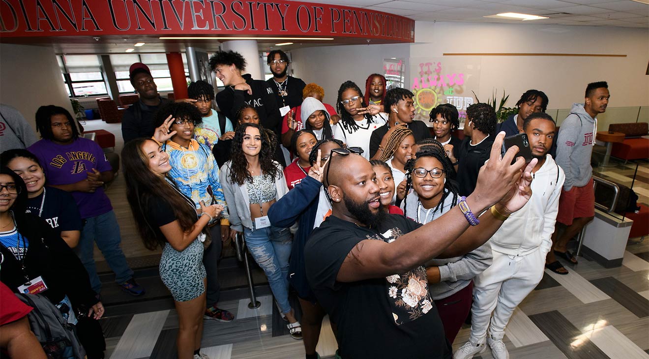 A young man with more than 20 younger people standing behind him holds up his phone facing himself and the crowd to take a selfie. They are in a large, lobby-type space with an “Indiana University of Pennsylvania” sign in red near the ceiling and a carpet with long rectangles in shades of gray.