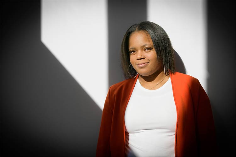 A young woman wearing a fitted white shirt and a red open sweater stands in front of a white wall and smiles slightly in the light and shadows created by sunlight coming through the windows of a building.