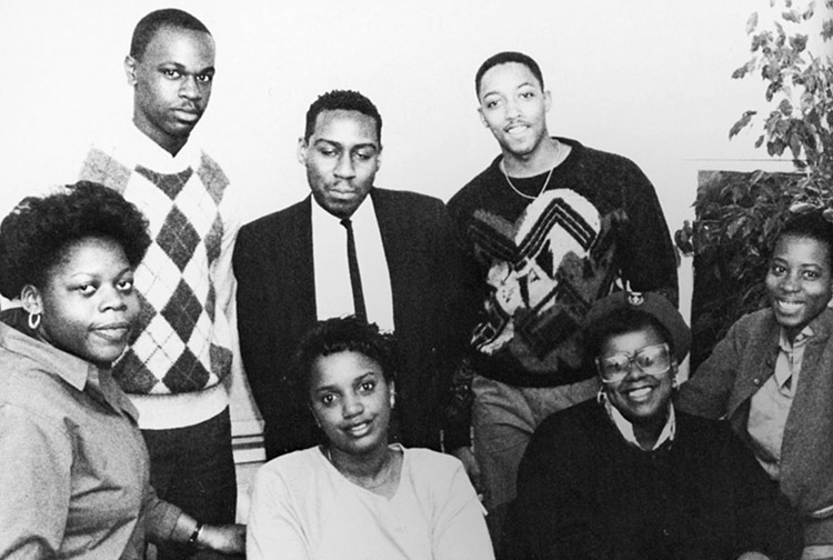 Black-and-white, posed photograph of seven young people in business casual clothing, with a plant in the background on the right. Four women sit in front, and three men stand behind them.