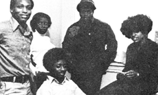 Black-and-white, posed photo of five young people in an office setting with a window behind them. Three are standing, one is seated at a desk, and the other is sitting on the desk.