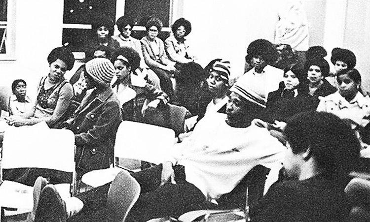 In this black-and-white photograph, about 20 young people in casual clothing, some wearing knit winter hats, sit in a classroom. Most are looking in different directions; some are talking amongst themselves. Three windows in back show it is dark outside.