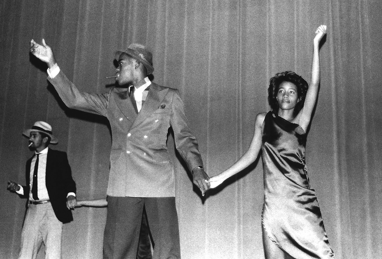 In the foreground of this black-and-white photo, a young man wearing a suit and fedora dances with a young woman in a silky, one-shouldered dress. They are holding hands, both facing the camera, and have their opposite hands raised. Another dancing couple is partly visible behind them.