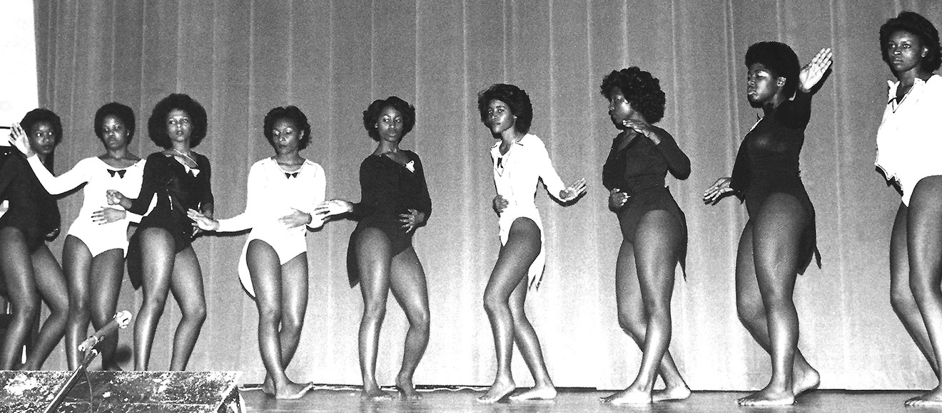 In this black-and-white photo, nine young women dance in a row on a stage, wearing either black or white leotards. Both sides of the row are turned to its center.