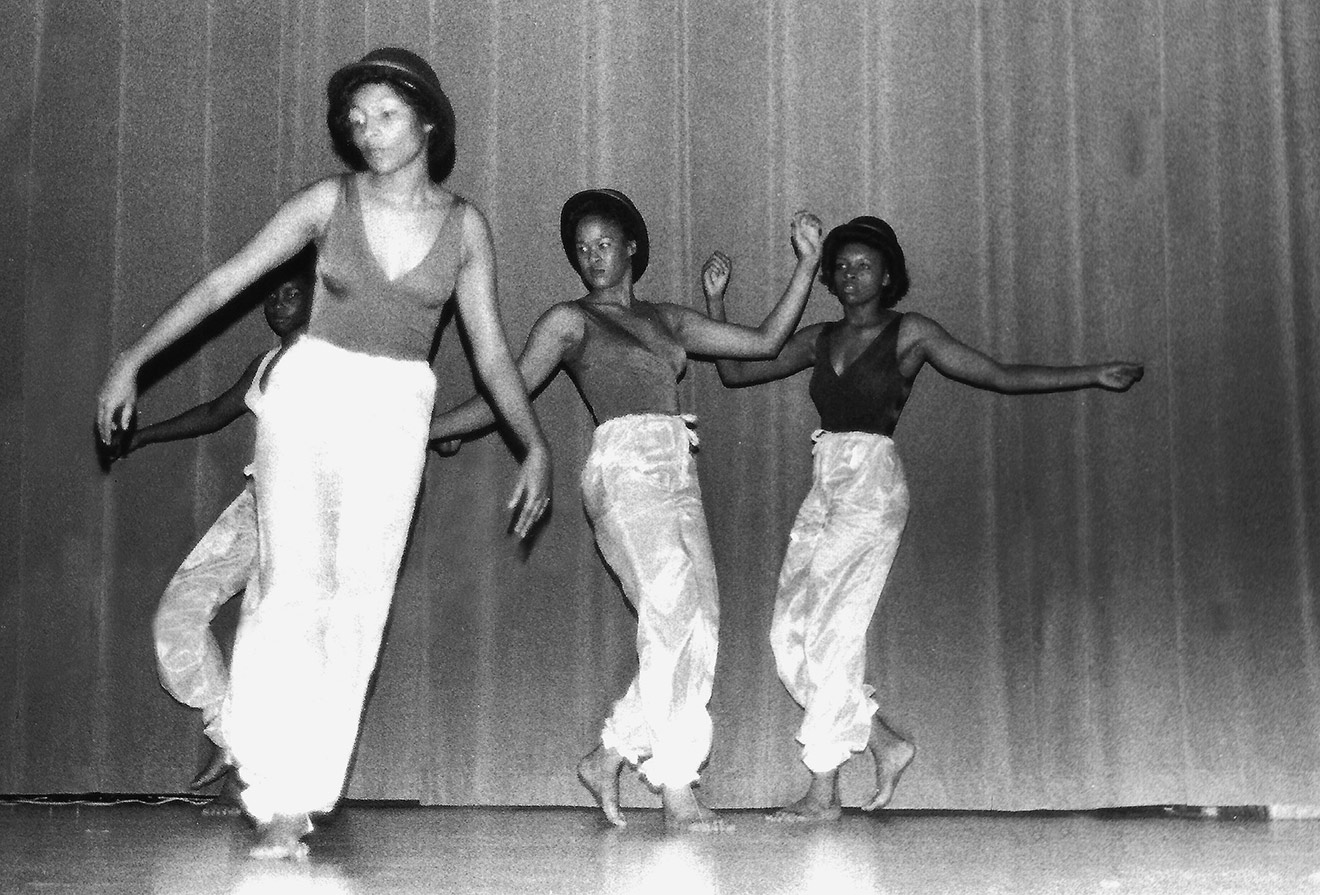 In this black-and-white photo, three young women and more in the background and to the side are dancing on a stage wearing dark, sleeveless, V-neck leotards and baggy light-colored pants that gather at the bottom.