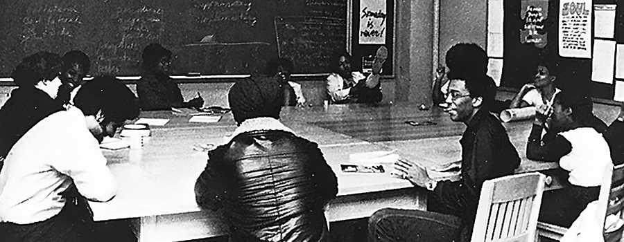 In this black-and-white photo, 12 people meet around four tables that are pushed together to form one large table in a classroom-type setting. In the background on the back wall is a chalkboard with writing and on a side wall is a bulletin board with posters.