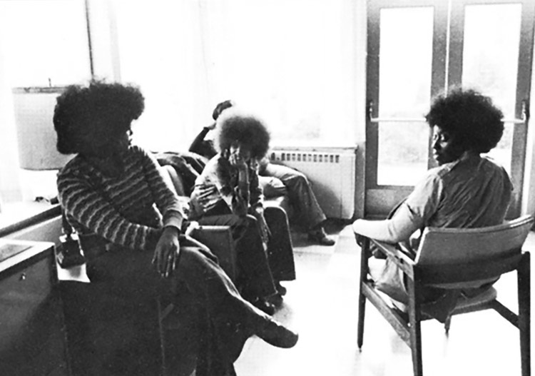 In this black-and-white photograph, three young women sit and talk in a lounge. Another person whose face is not visible is sitting behind them. One woman has her head turned to look at the camera.